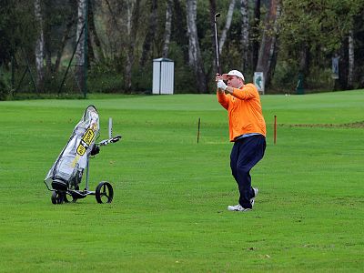 A golfer in an orange sweater swings on a green course with a golf cart nearby, surrounded by trees and an overcast sky.