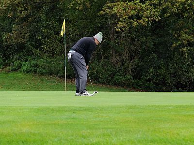 A golfer putts on a green golf course, wearing a black sweater and gray pants. The flagstick stands behind him, with trees in the background.