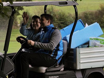 Two people ride in a golf cart. The passenger waves happily while the driver navigates. Supplies are visible in the back of the cart.