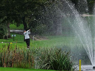 A golfer swings near a large fountain on a green course, with water spraying into the air and trees in the background.