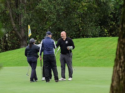 Four people stand on a lush golf course green, smiling and shaking hands. Trees and a golf flag are visible in the background, indicating a friendly game.