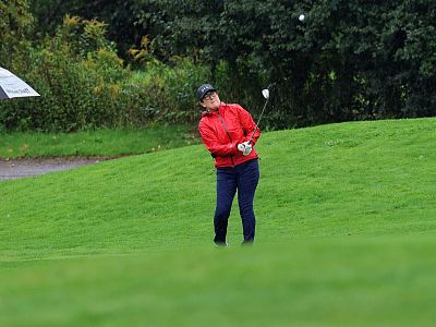 Golfer in red jacket swings club on green course, surrounded by trees; umbrella visible on left.