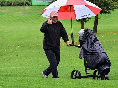 Person with red and white umbrella pushing a covered golf trolley on a rainy golf course, giving a thumbs up.