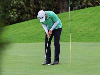 Golfer in green shirt and cap putts on golf course green, surrounded by lush greenery and trees; focusing on the ball near the hole.