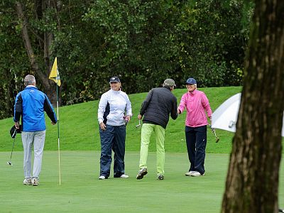 Four golfers stand on a green lawn with an umbrella nearby, indicating a rainy day. They are dressed in casual sportswear, gathered near a golf flag.