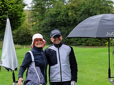 A couple in golf attire posing on a green with a golf umbrella and trees in the background, both smiling widely.