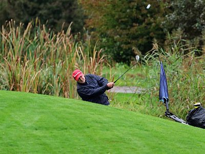 Golfer in red cap hitting a shot from thick grass on a green course, with tall grass in the background and a nearby golf bag with an umbrella.