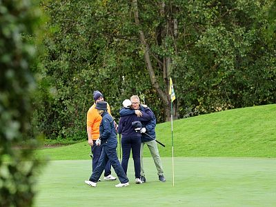 Four people on a golf course celebrate near a flagstick on a green, surrounded by trees and greenery.