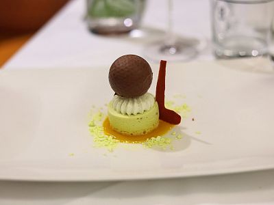 A dessert on a plate with a green base, topped with a chocolate sphere, white cream, and red garnish on a white tablecloth background.