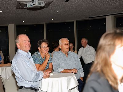 Group of people in semi-formal attire at an indoor event, standing around a table with a white cloth, with dim lighting and more guests visible in the background.