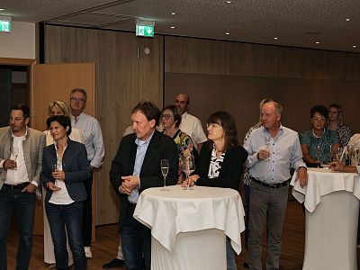 People at a corporate gathering, standing around tall tables with drinks, in a wood-paneled event space. They're engaged in conversation and dressed smart casual.