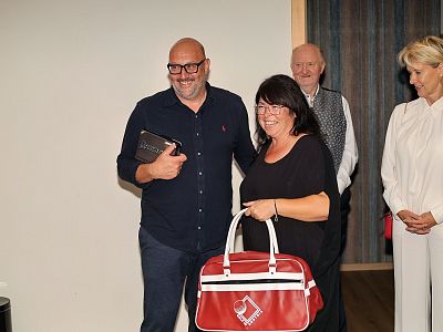 A man and woman smiling, holding a gift bag and a tablet, with two smiling people in the background, in a casual indoor setting.