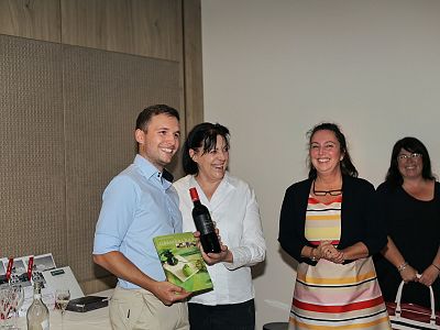 Four people in a room; two exchanging a wine bottle, smiling, while two others smile in the background, one wearing a striped dress.