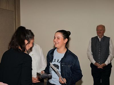 Two people exchanging an award certificate, smiling warmly, with a small audience in the background observing the scene in a room setting.