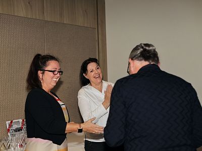 Three people in a friendly conversation in an office. Two women smiling while talking to a man, with a wooden wall in the background.