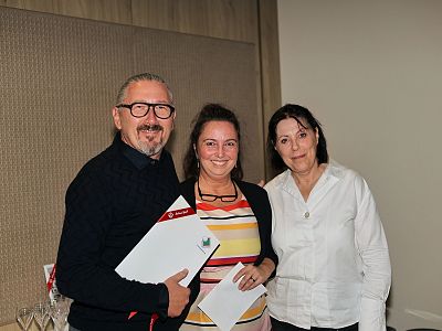 Drei Personen posieren lächelnd in einem Raum. Die Person links hält ein Dokument, während die anderen beiden in die Kamera schauen.
