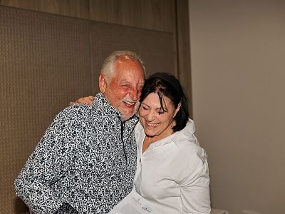 A joyful elderly couple laughing and embracing each other in a warmly lit room. The man is wearing a patterned shirt and the woman a white blouse.