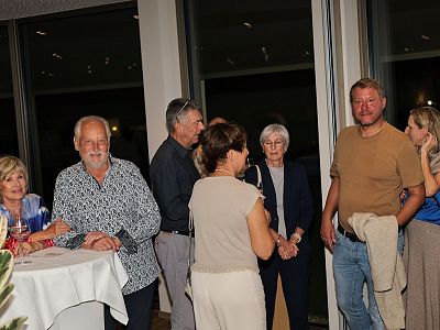 A group of people standing in a room by large windows. Some are talking, others are standing near a table. The atmosphere appears to be relaxed and social.
