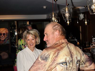 Eine Frau und ein Mann stehen lächelnd in einem gedämpft beleuchteten Raum. Der Mann trägt ein Hemd mit einem Löwenmotiv. Im Hintergrund sind Lampen zu sehen.