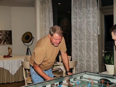 Zwei Männer spielen Tischfußball in einem gemütlichen Raum mit dekorativen Vorhängen. Im Hintergrund steht ein Tisch mit Tischdecke und Dekorationen.