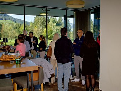 A group of people talking in a modern room with large windows displaying a view of trees and hills outside.