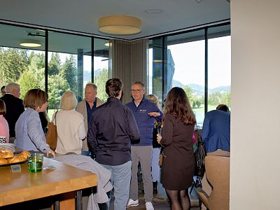 People networking in a bright room with large windows, engaging in conversation and enjoying refreshments. Trees and greenery are visible outside.