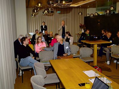 People in a modern conference room engaging in discussion and note-taking. The room has wooden tables and stylish lighting fixtures.