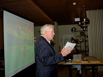 A man in a suit speaks in a meeting room, holding papers. A screen with colorful visuals is visible beside him. Modern interior with spherical lights.