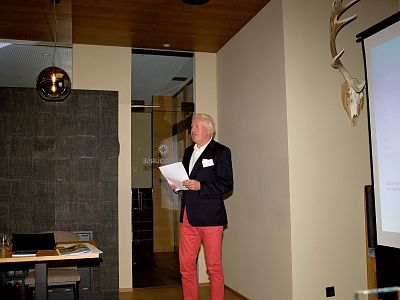 A man in red pants and a blazer gives a presentation in a modern room. A projector screen and a mounted deer head are visible in the background.