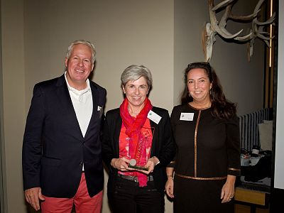 Three people standing together inside at an event, two women and a man. One wears a red scarf, another a black dress, and the man a navy blazer and pink pants.