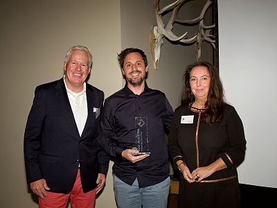 Three people at an award ceremony, with the person in the center holding a trophy. They are dressed formally, and decorations are visible in the background.