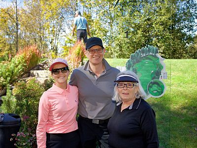 Three people outdoors in casual wear, smiling in a sunny garden. A person stands on a hill behind them, with trees and grass in the background.
