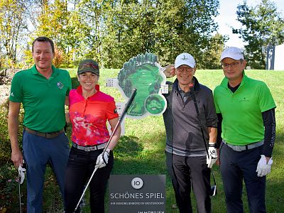 Four individuals posing with golf clubs, two wearing caps and all in sporty attire, stand on lush grass with trees in the background. They smile for the camera.