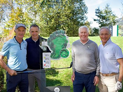 Four men stand together on a sunny golf course. One holds a golf club, another holds a clear award. Trees and grass surround them. They're smiling.