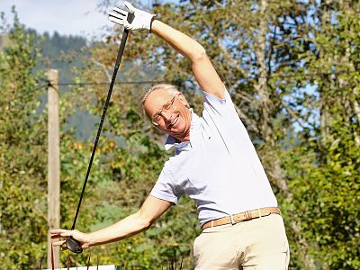 A man is stretching with a golf club outdoors, surrounded by trees and hills. He's wearing a light shirt and beige pants, looking cheerful under a sunny sky.