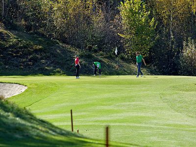 Golfers play on a lush green golf course, surrounded by autumn-colored trees and sunlight creating long shadows. The scene is calm and picturesque.