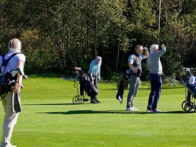 Four golfers stand on a lush green course, two holding clubs, two watching intently. Trees in the background under a clear, sunny sky.