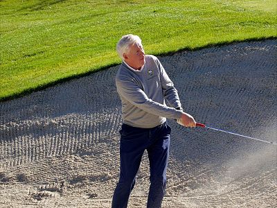 A golfer in a sand bunker wearing a grey sweater and blue pants, mid-swing to hit a golf ball. The grass around the bunker is bright green.