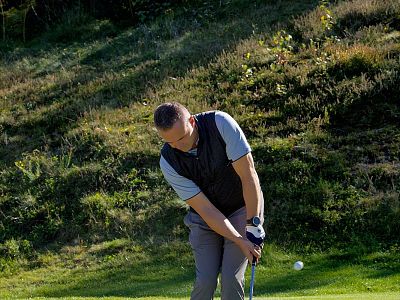 A man in casual attire is preparing to hit a golf ball on a grassy area, surrounded by greenery and sunlight.