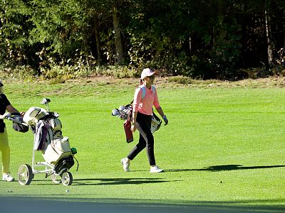 Two golfers on a green course; one pulls a cart, the other carries clubs, under a sunny blue sky and near a forest edge.