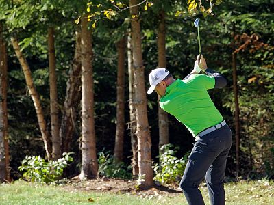 Golfer in bright green shirt swings club in a forest setting, surrounded by tall trees and greenery on a sunny day.