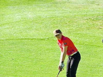 A woman wearing a red shirt and cap is swinging a golf club on a green course. She is concentrating on her stance. The day is sunny and bright.