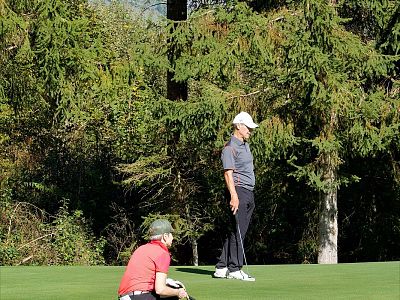Two people on a golf course; one standing with a club, the other crouching. Trees in the background add a natural green setting.