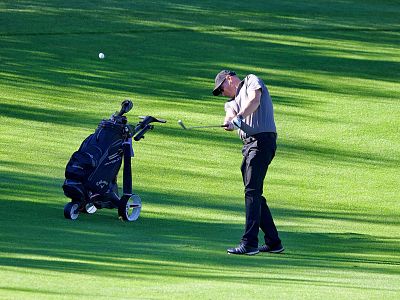 A golfer swings his club on a green course, shadows on grass, with a golf cart beside him.
