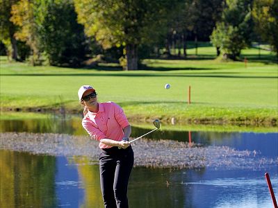 A golfer wearing a pink shirt is swinging a golf club near a small water hazard on a golf course. The sky is clear, with green trees in the background.