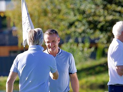 Two men on a golf course having a conversation. One is smiling at the camera, the other has his back turned. Both wear light shirts, under clear blue skies.