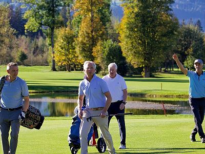 Four individuals walking on a sunny golf course, holding clubs. Greenery and a water feature in the background, signaling a pleasant day outdoors.