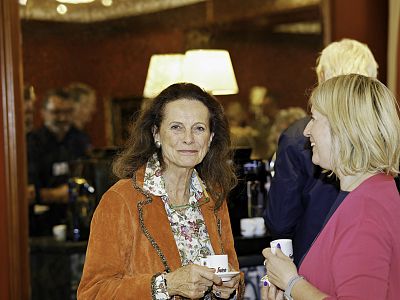 Two women are conversing, each holding a cup, in a warmly lit room with people and lamps in the background, creating a welcoming atmosphere.