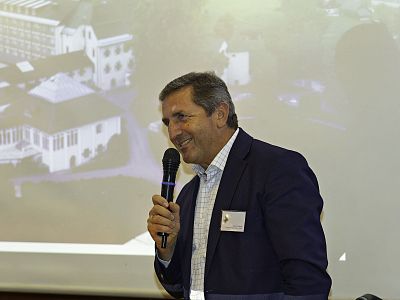 A person in a dark suit holding a microphone in front of a projected image at a conference, appearing engaged and smiling.