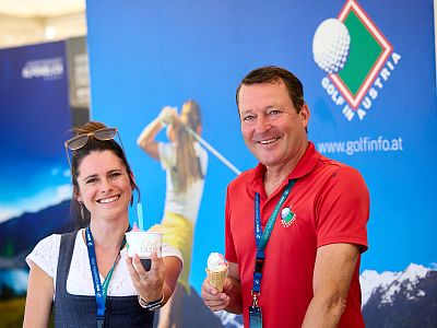 Two smiling people eat ice cream in front of a golf-themed background at an event.